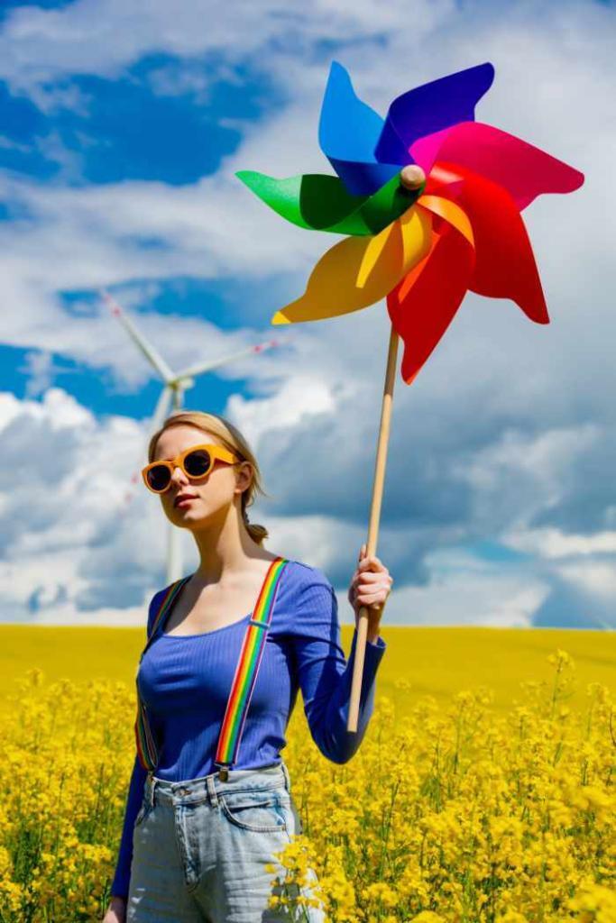 Beautiful female in rainbow suspenders, blue long-sleeved top and blue jeans stands in a field full of tall yellow flowers. In her left hand, she is holding a large rainbow pinwheel with a long wooden handle. She is wearing orange sunglasses. Behind her is a white wind turbine. Above her is a blue sky smattered with puffy white clouds.