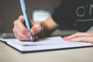 A person writing in a notebook with a pen. We can only see their arms. They are wearing a dark t-shirt.