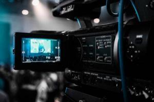 A broadcast video camera viewed from the back left. It is black with a flip-out LCD view screen that has a person showing.