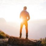 A man stands on a mountaintop looking off into the distance toward the sunset. We see him from behind as he takes in the vista.