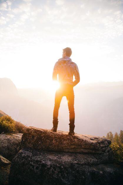 A man stands on a mountaintop looking off into the distance toward the sunset. We see him from behind as he takes in the vista.