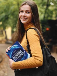 A young, pretty woman with long brown hair is on her journey to life mastery. She stands with her face turned toward the camera. She is wearing a colorful long sleeved top with a black backpack slung over her left shoulder. In her arms she carries a copy of Volume #1 of The Spin Positive Master Series. It is a blue book with a cover featuring swirling energy. The words "Spin Positive Master Series The Universe's Guide to Mastering Your Power Volume One Blue Level" are visible. Behind the woman is a serene parklike setting with trees and various structures. The weather is pleasant and sunny.