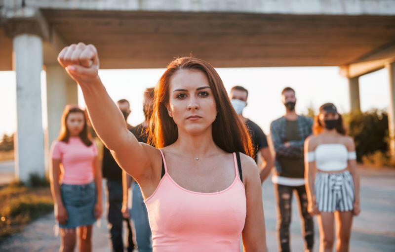 A group of young people standing under a bridge. In the back are four boys and two girls. The girls are on each side. The boys in the middle. They are arranged from shortest to tallest to the middle then tallest to shortest. Making an arc of people in the background. In the front is a young woman wearing a white spaghetti strap top. She has her right arm raised in a fist. She has a determined expression on her face.