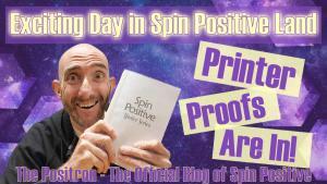 A graphic with a blue background depicting the universe with stars and nebula present. Along the sides, large faceted blue hexagons are found. Along the top the words Exciting Day in Spin Positive Land. To the center right of the picture are the words Printer Proofs Are In. To the center left is a picture of handsome curator of Spin Positive Mike Garrett. He is smiling and is shot from the mid torso up. He is wearing a black sweatshirt and is holding a copy of Spin Positive Master Series Volume #1 in his hand. The book is a printer proof copy so it is missing its cover. Along the bottom are the words The Positron - The Official Blog of Spin Positive