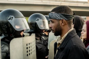 A black man with a stubble beard wears a bandana and a dark jacket while facing off against a line of police in riot gear.