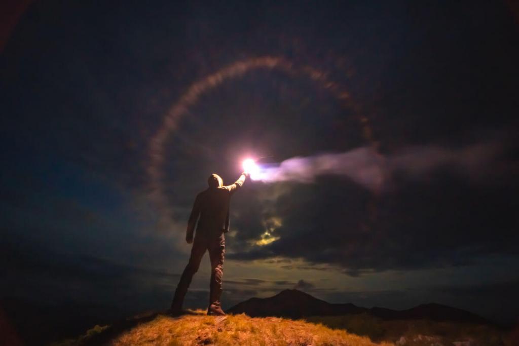 A man in a hoodie and jeans stands on top of a mountain. It is dark outside, but he holds in his right hand a lit flare burning bright and held high. There is a halo of light colors around the bright center. A mountain stands proud in the distance. Dark clouds pepper the sky.