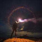 A man in a hoodie and jeans stands on top of a mountain. It is dark outside, but he holds in his right hand a lit flare burning bright and held high. There is a halo of light colors around the bright center. A mountain stands proud in the distance. Dark clouds pepper the sky.