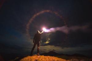 A man in a hoodie and jeans stands on top of a mountain. It is dark outside, but he holds in his right hand a lit flare burning bright and held high. There is a halo of light colors around the bright center. A mountain stands proud in the distance. Dark clouds pepper the sky.