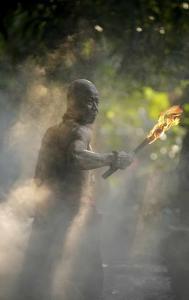 a man with bronze skin is leading the way down a smoky path. He is bald, wearing ceremonial battle garb, and carrying a flaming torch in his right hand. There are tree branches above the path. Sun sprinkles in from ahead afar.