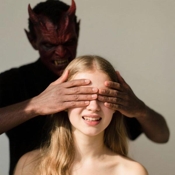 A demon-looking dark worker with red skin, horns, and fangs stands behind a seated, young white girl. She appears to be naked from what we can see. The dark worker has his hands over the girls eyes so she cannot see.
