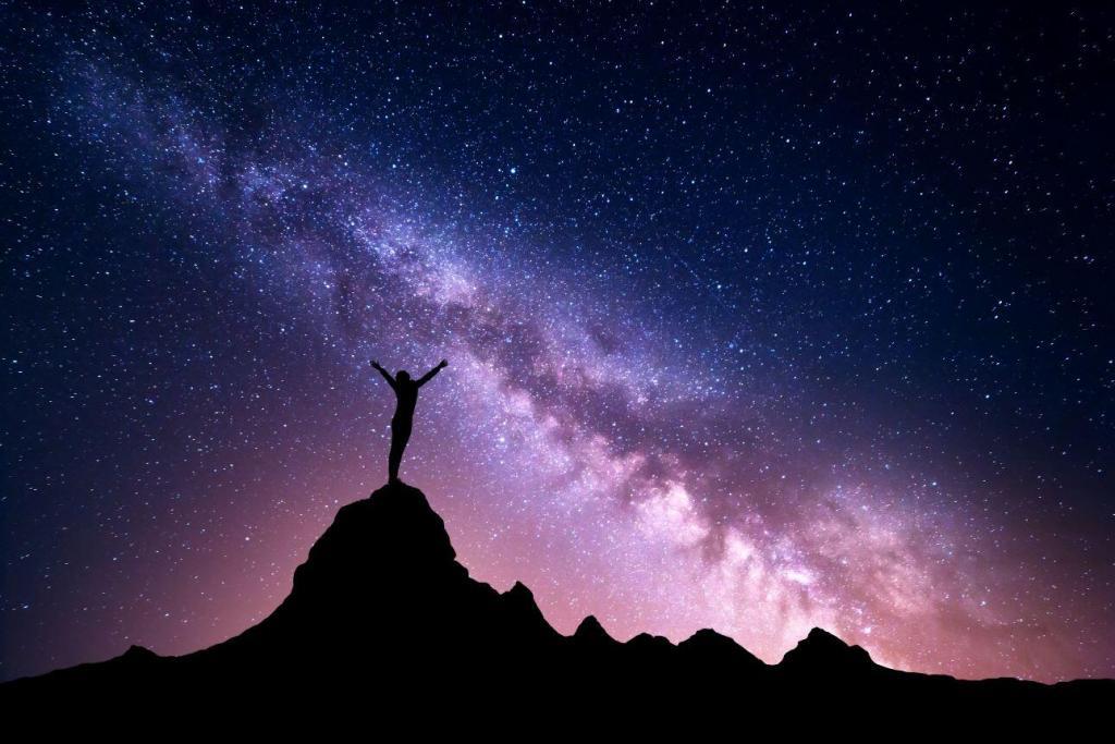 A person stands on top of silhouetted mountain against a blue tinged night sky. The stars and Milky Way are visible in the sky. The person has their arms raised up in awe and adoration of the beautiful scene.