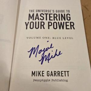 Volume #1 of the Spin Positive Master Series is open to its title page. It has the normal verbiage. "The Universe's Guide to Mastering Your Power, Volume #1 Blue Level", a large space and then the words "Mike Garrett, Penny Apple Publishing." In the space on this copy, Mojoe Mike has signed it in blue ink, and while signing he set powerful intentions over the volume to boost the readers revolution for life mastery.