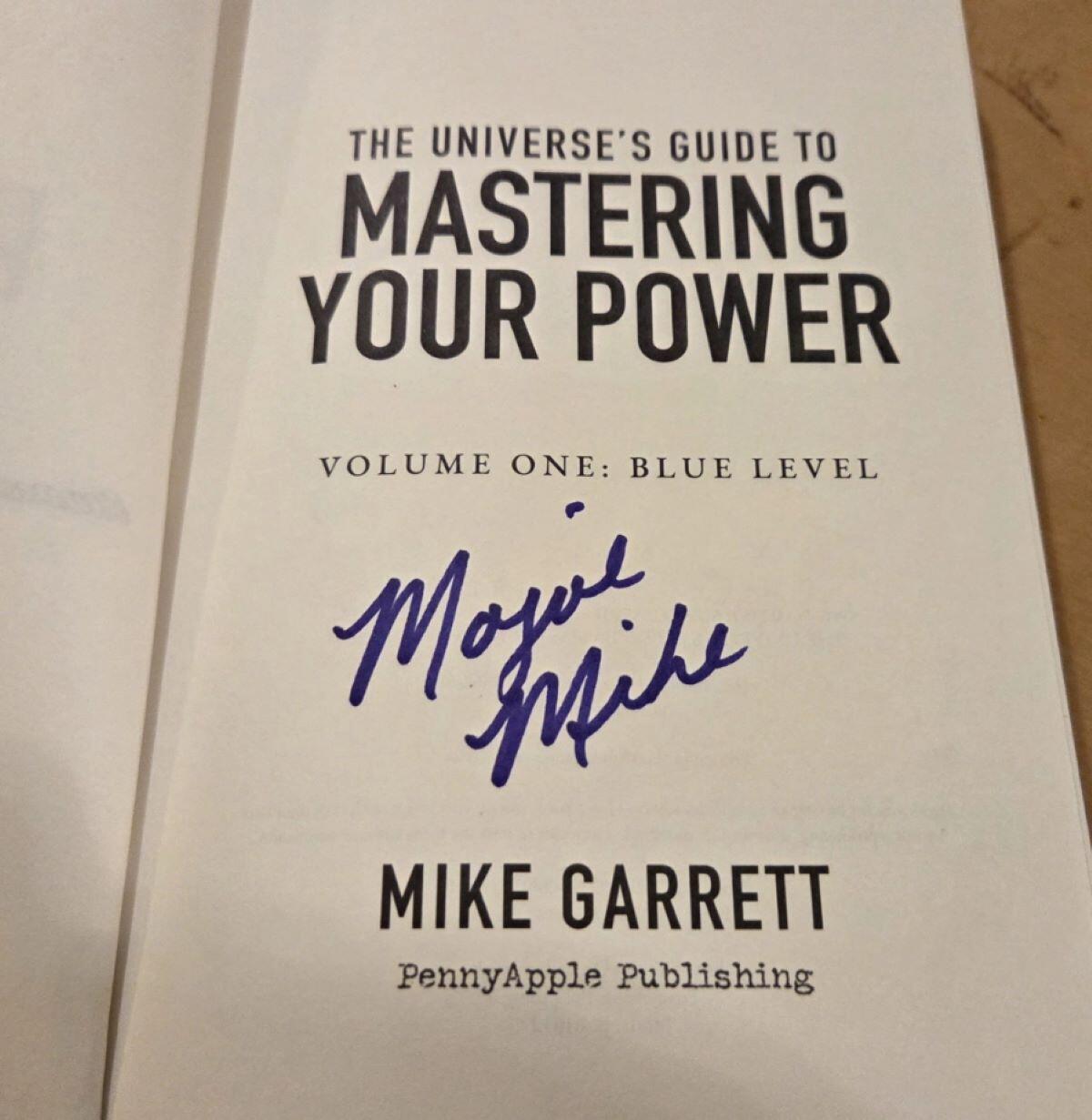Volume #1 of the Spin Positive Master Series is open to its title page. It has the normal verbiage. "The Universe's Guide to Mastering Your Power, Volume #1 Blue Level", a large space and then the words "Mike Garrett, Penny Apple Publishing." In the space on this copy, Mojoe Mike has signed it in blue ink, and while signing he set powerful intentions over the volume to boost the readers revolution for life mastery.