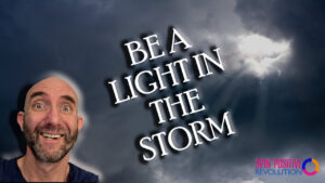 A dark, grey, stormy, cloud-filled sky lets a ray of sunlight peek in. In the lower left corner is a headshot of Mojoe Mike outlined in a yellow glow. He is smiling with a fun look on his face. Opposite in the lower right hand corner is the Spin Positive Revolution logo with the tri-arrow graphic. The words "Be a Light in the Storm" are bolded in white at the center of the image.